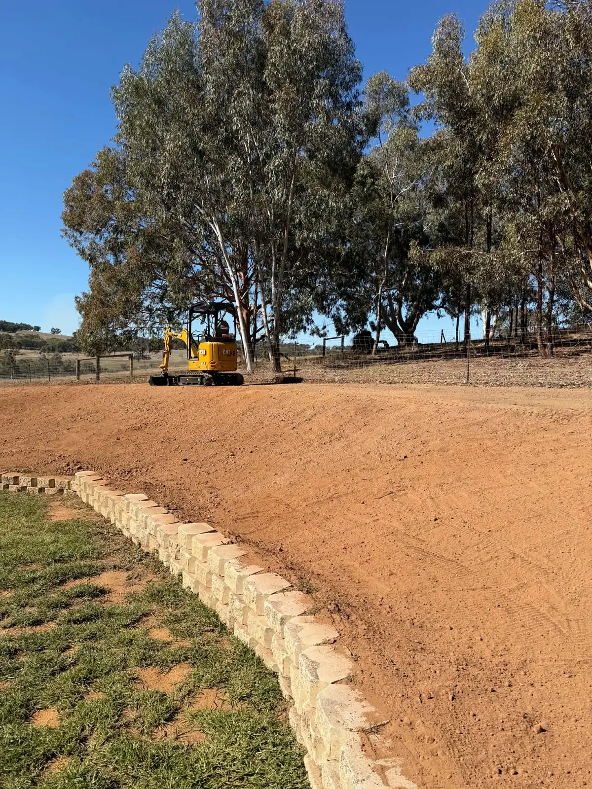 Yellow mini excavator at the crest of a freshly graded red dirt batter above a limestone block retaining wall