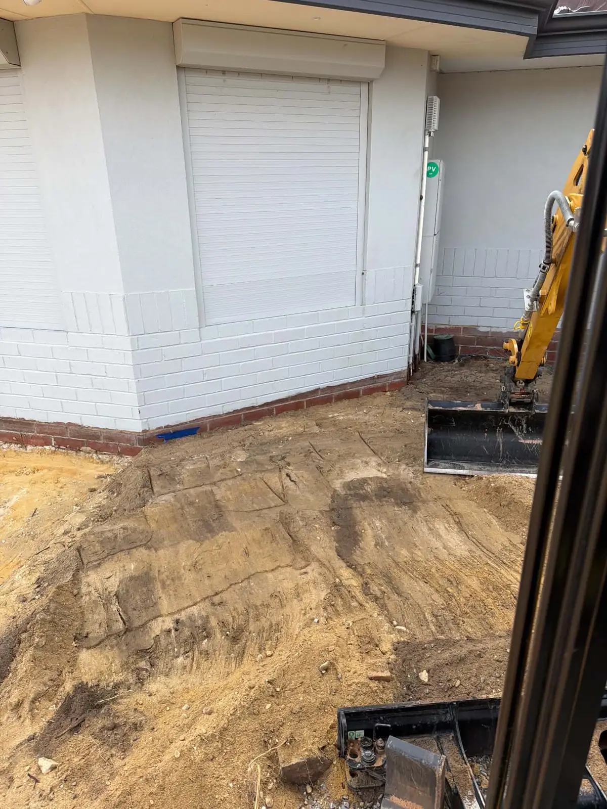 View from inside the excavator cab of the boom and bucket digging beside a white brick house