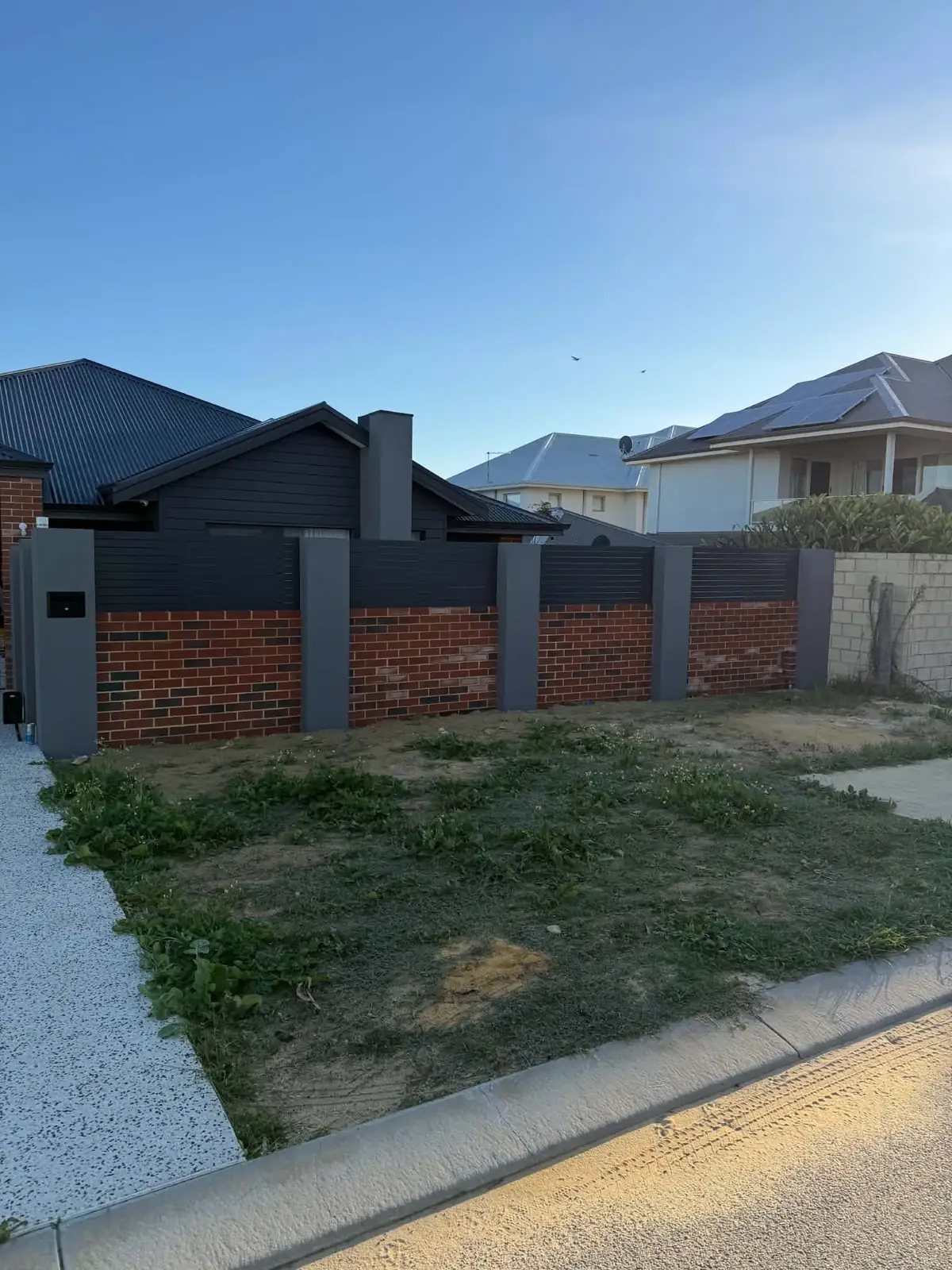 A vacant lot of weeds and loose sand beside a kerb and exposed aggregate driveway, in front of a brick and slat fence