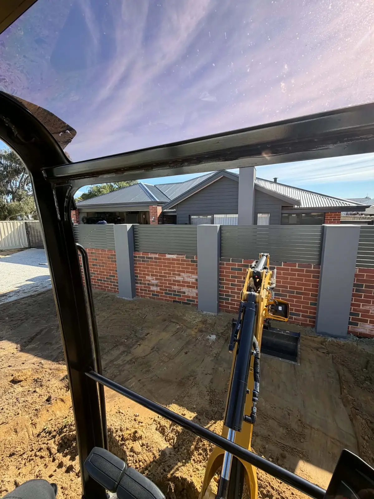 View from the excavator cab of the boom and bucket working over dug up sand in front of a brick and slat fence